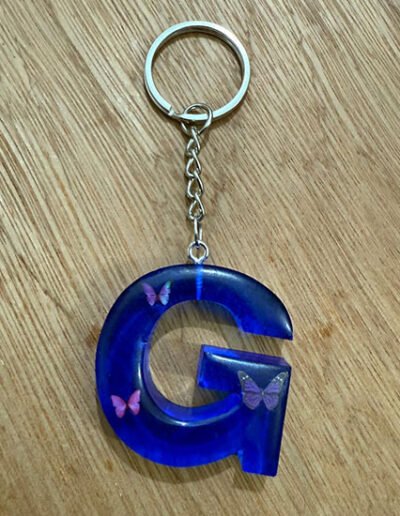 Blue resin keychain shaped like the letter G with embedded butterfly decorations on a wooden surface.