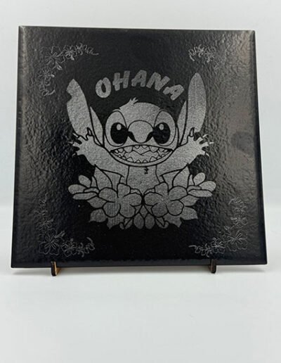 Black ceramic tile engraved with a cartoon character, floral motifs, and the word “Ohana”.