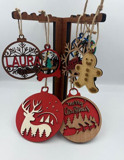 Assorted custom wooden and acrylic Christmas ornaments hanging on a stand, including reindeer, gingerbread man, name and “Merry Christmas” designs.